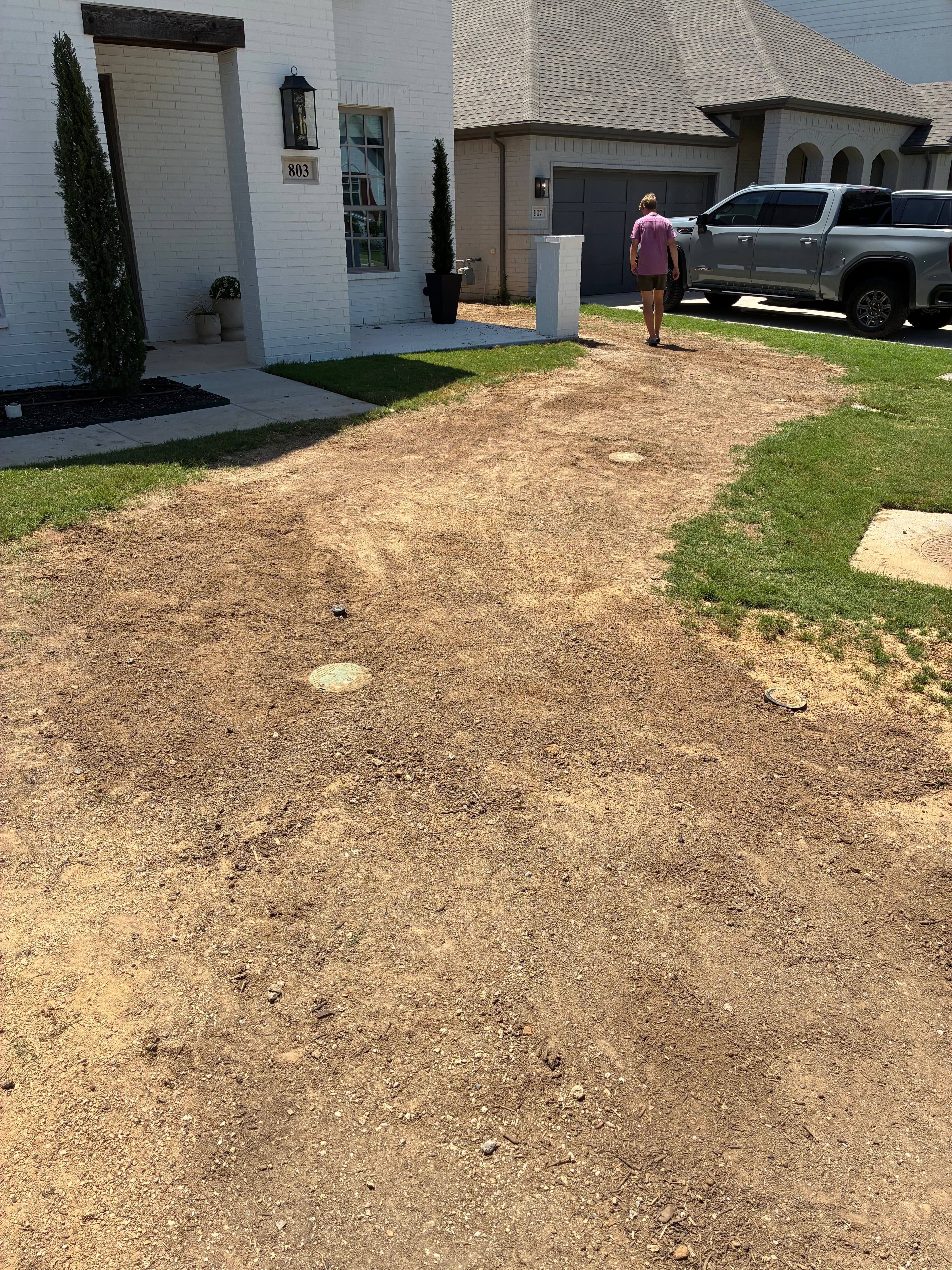 Man walking on a dirt driveway near a home and parked truck, with grass patches visible.