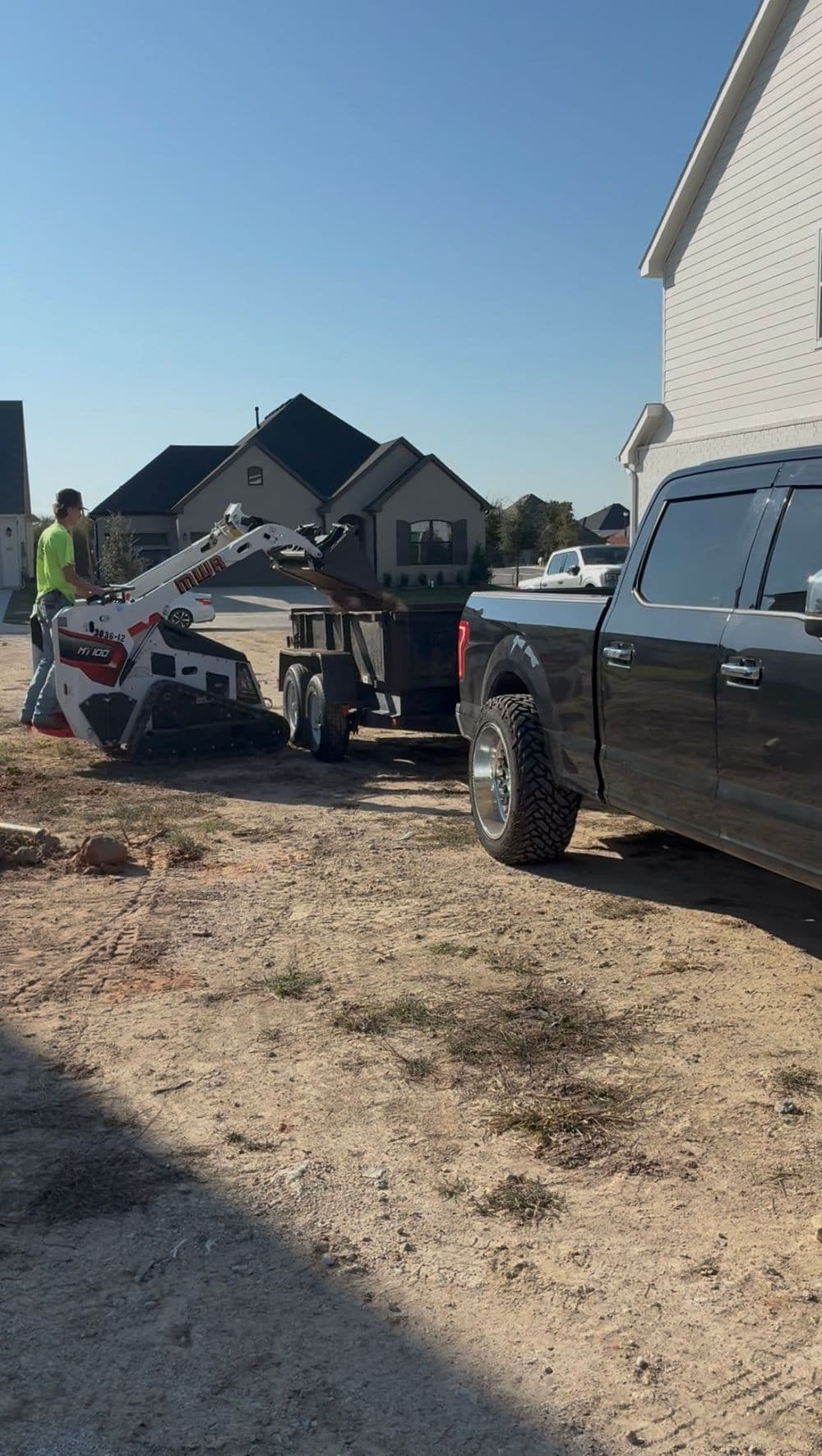 Construction worker operating a skid steer loader near a trailer and pickup truck.