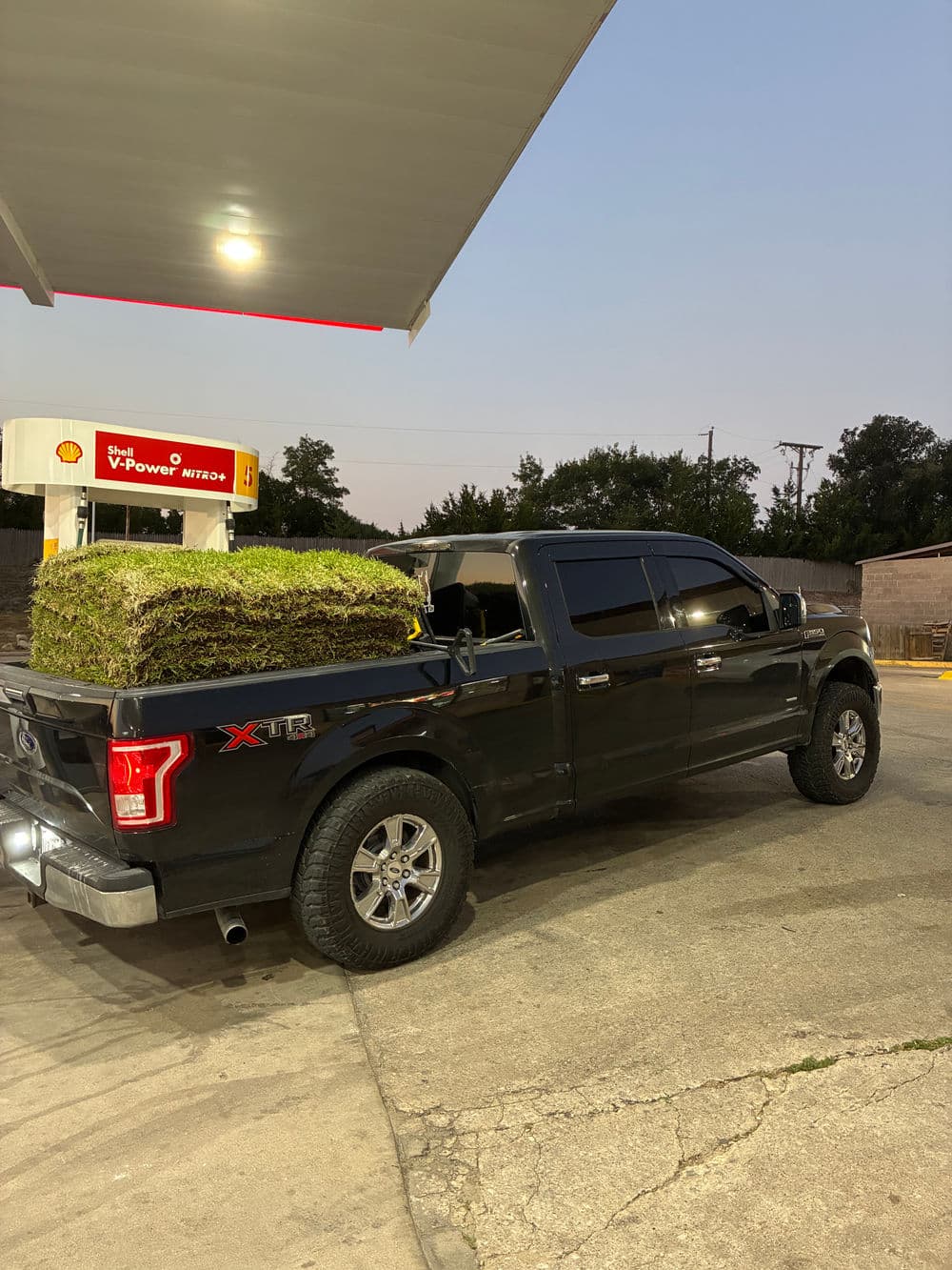 Black pickup truck with a grass sod pallet parked at a gas station at dusk.
