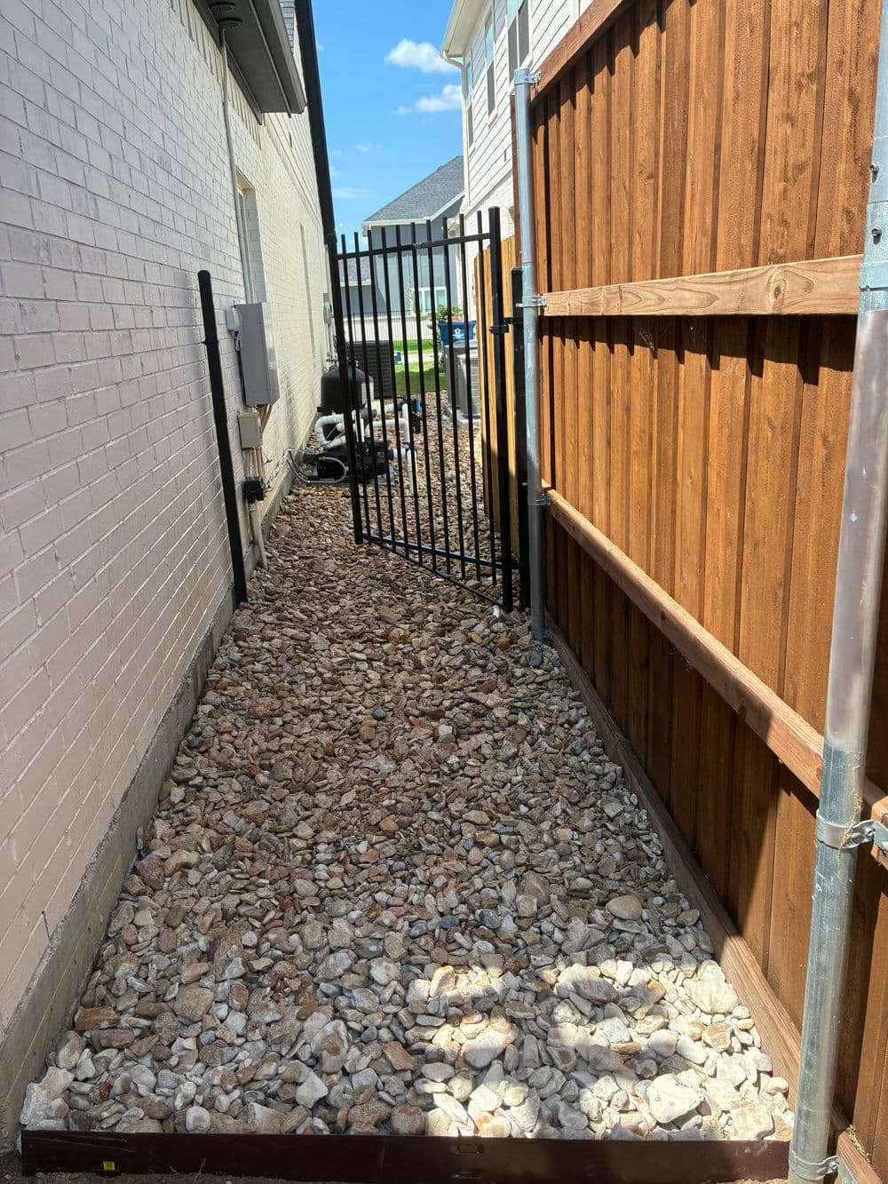 Narrow gravel pathway between two buildings, featuring a wooden fence and a gate.