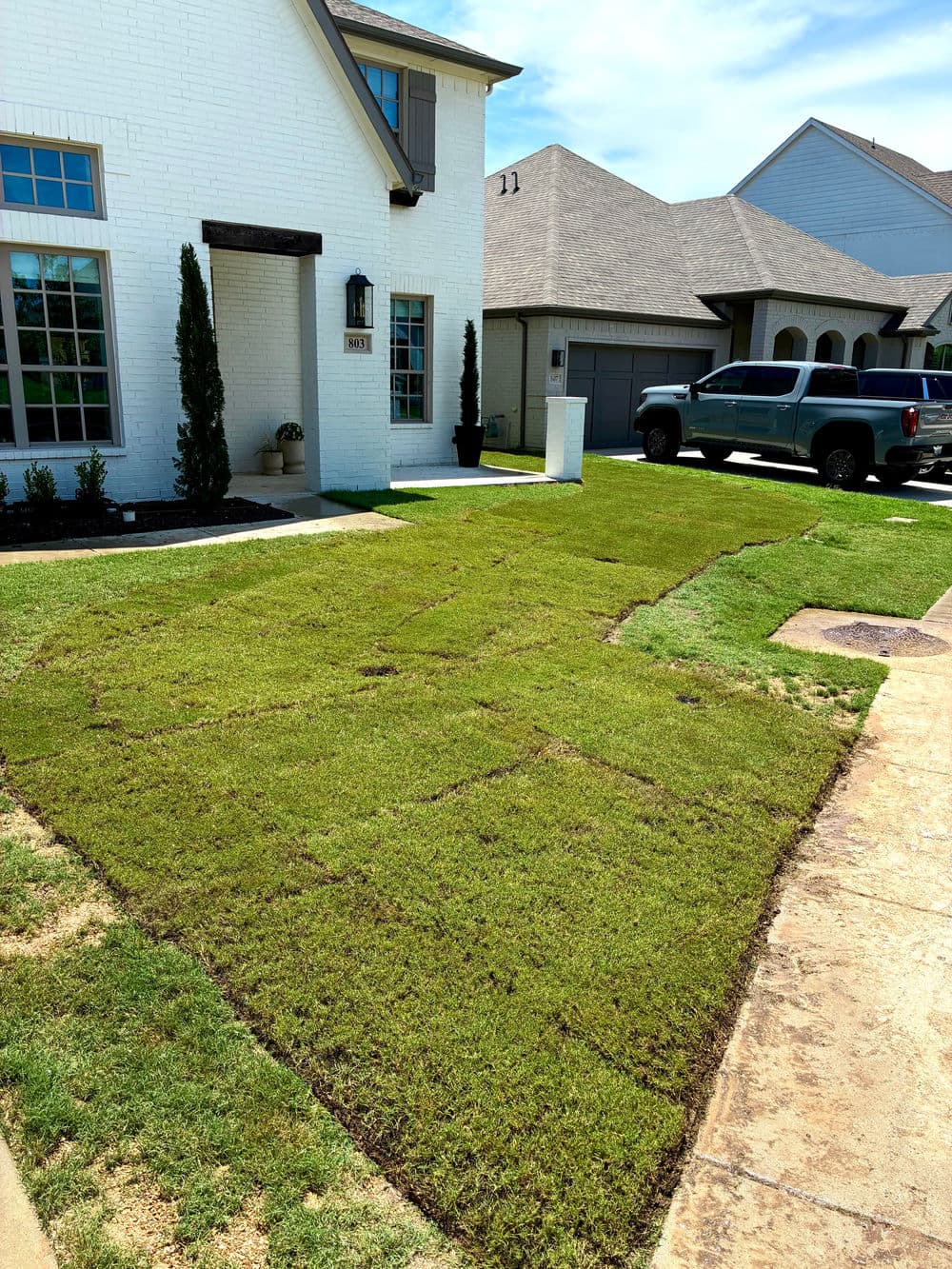 Newly installed sod lawn in front of a house, with unpaved sections and vehicles nearby.
