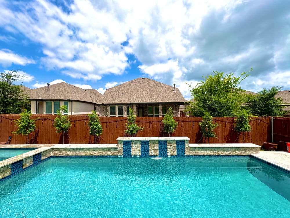 Bright outdoor swimming pool with a stone border, surrounded by trees and houses under a cloudy sky.