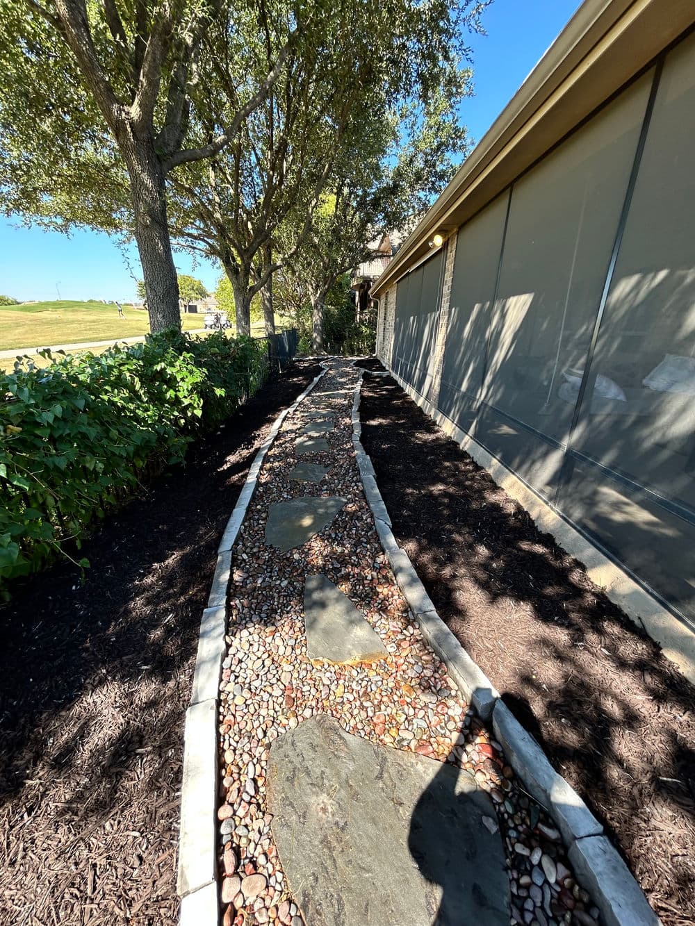 Stone pathway lined with pebbles and bordered by greenery next to a house.
