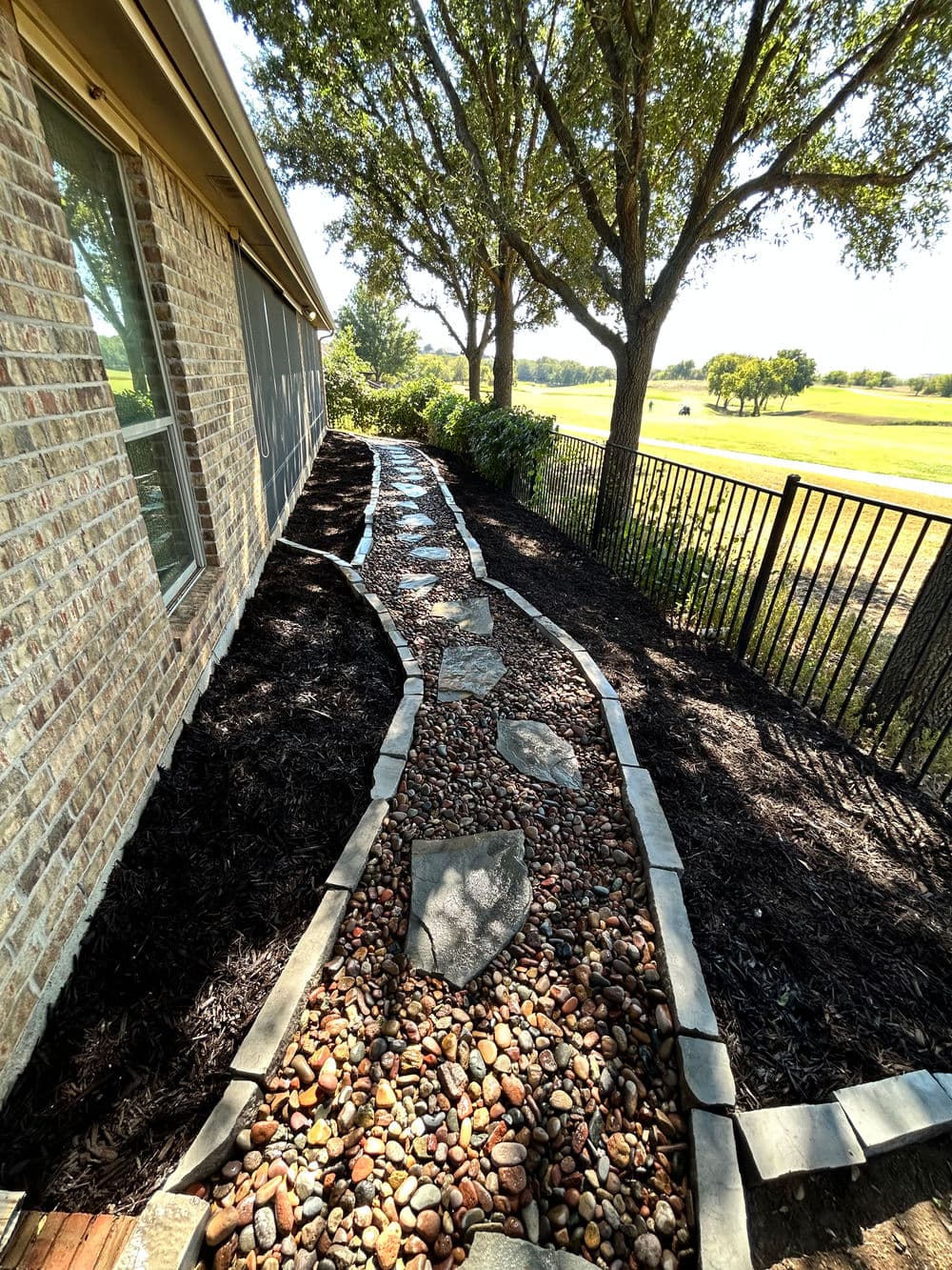 Landscaped pathway with pebbles and stepping stones beside a brick house and trees.