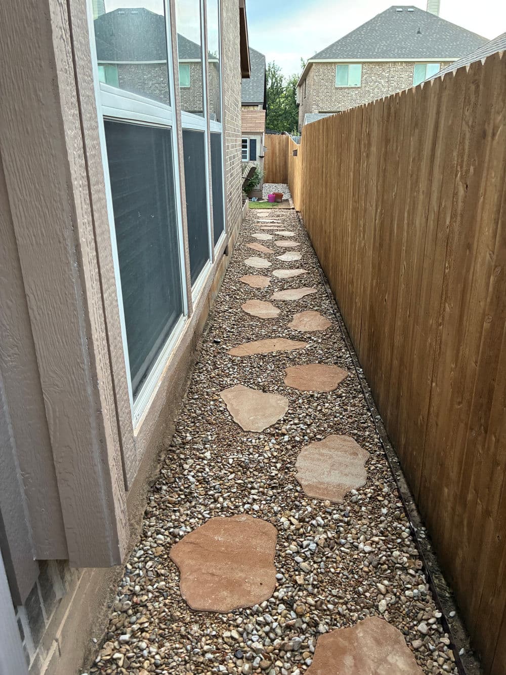 Narrow garden path with stone stepping stones and gravel, bordered by wooden fence.