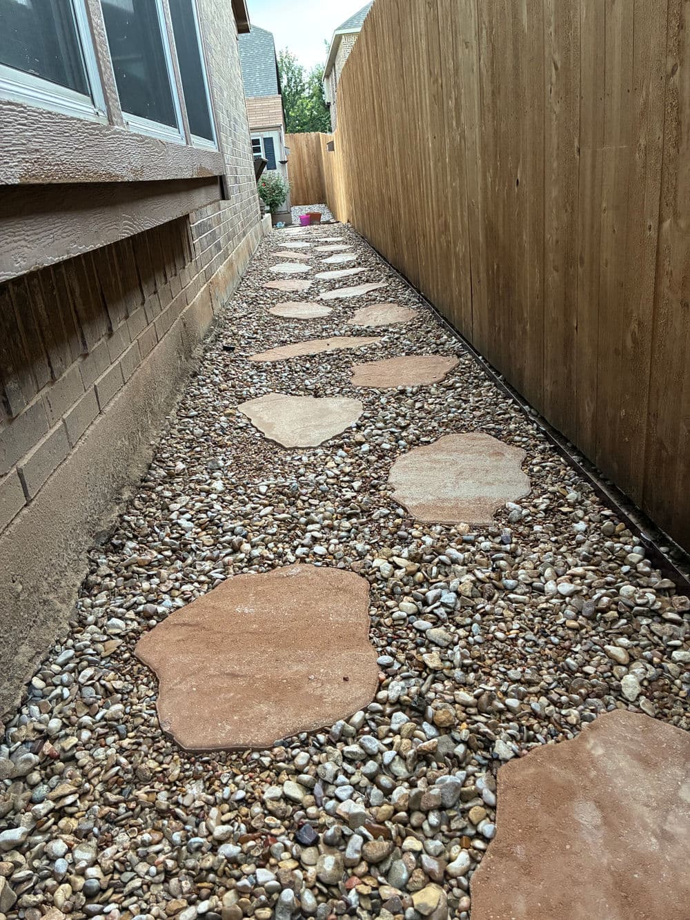 Garden path with stone stepping stones and gravel bordered by wooden fence and houses.