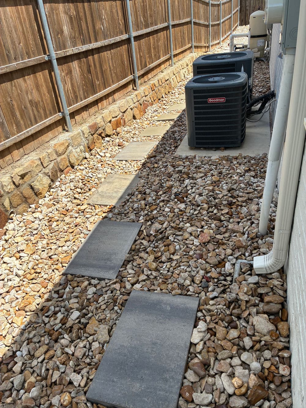 Stone pathway alongside air conditioning units in a gravel yard with wooden fence.