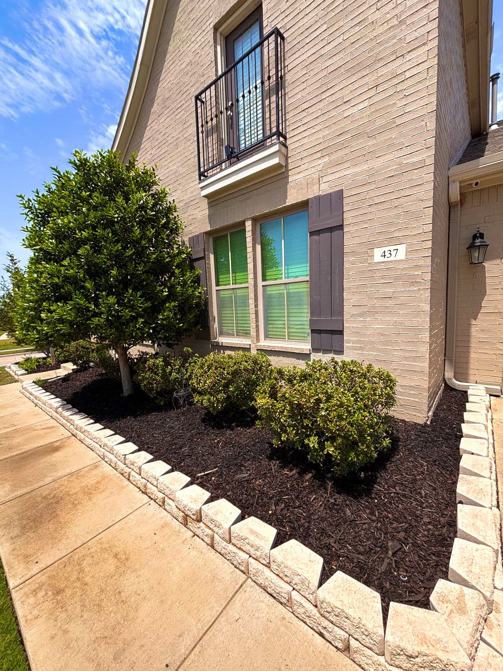 Well-maintained landscaping with shrubs and fresh mulch beside a brick house and sunny blue sky.