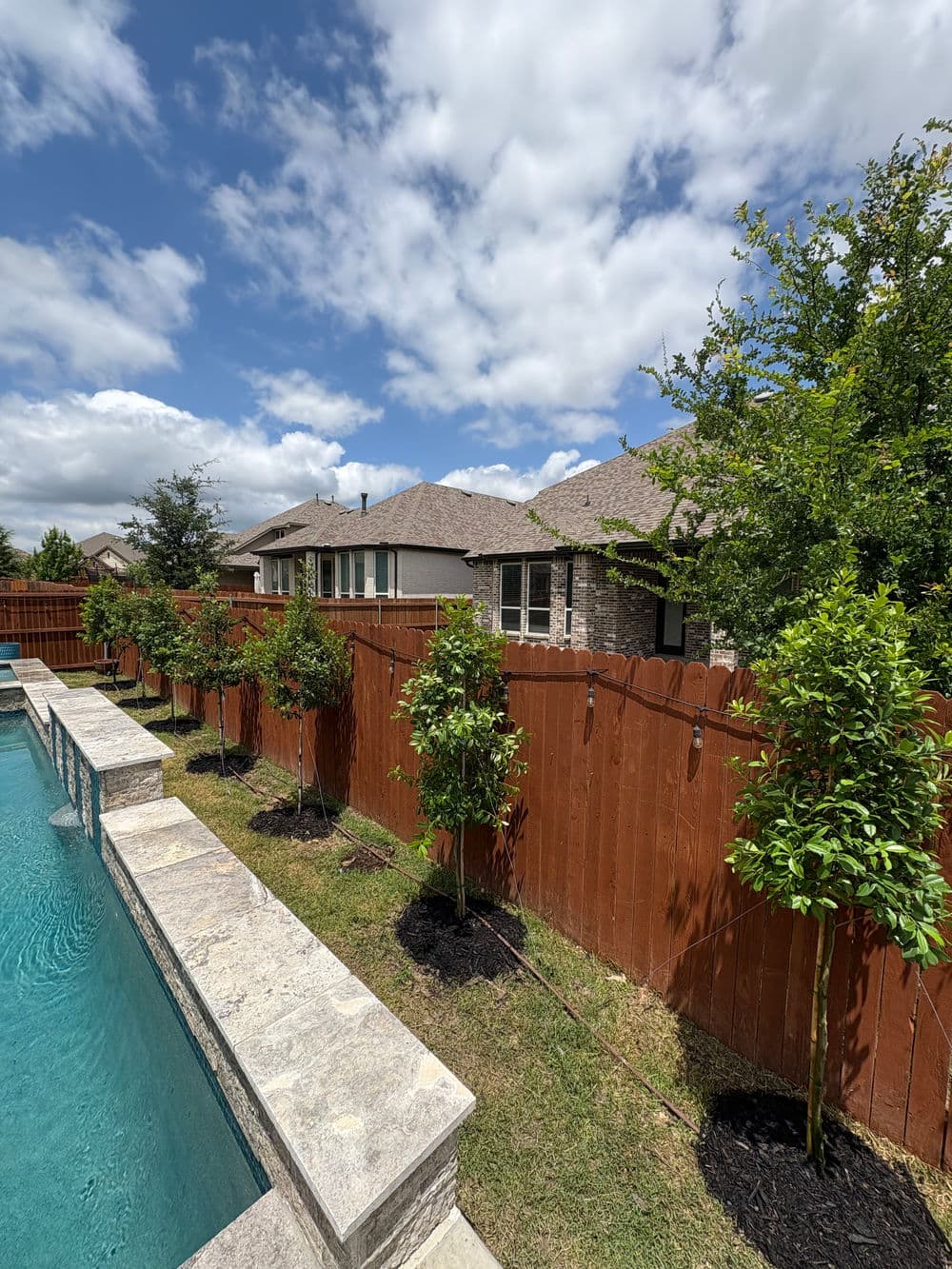 Backyard view featuring a pool, landscaped trees, and a wooden fence under a cloudy sky.